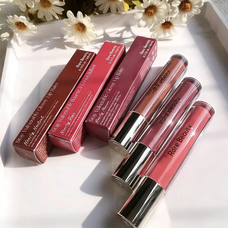 Rare Beauty Stay Vulnerable Glossy Lip Balm trio in neutral, rose, and mauve shades with boxes on a white tray.