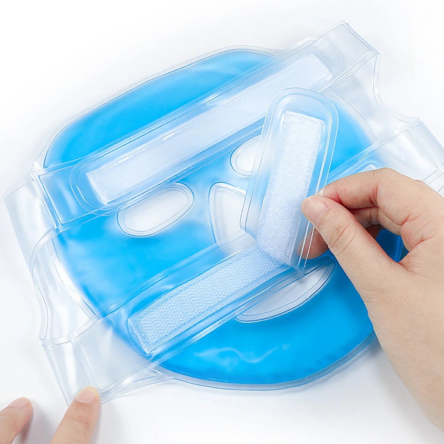Blue gel cooling face mask made of clear PVC with adjustable velcro straps, shown being secured by a hand.