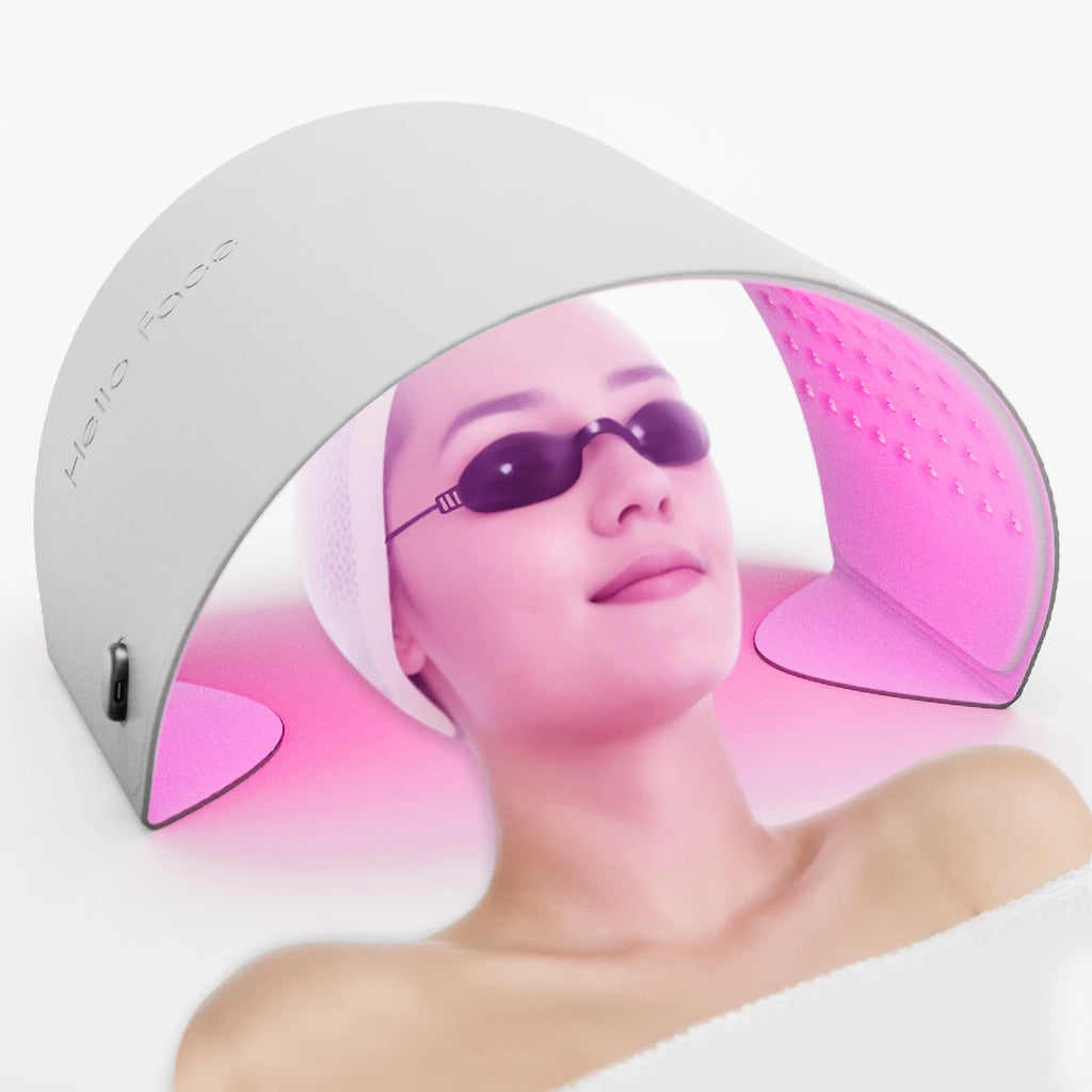 A woman wearing goggles lies under an arched, white Hello Face LED therapy device emitting a vibrant pink light.