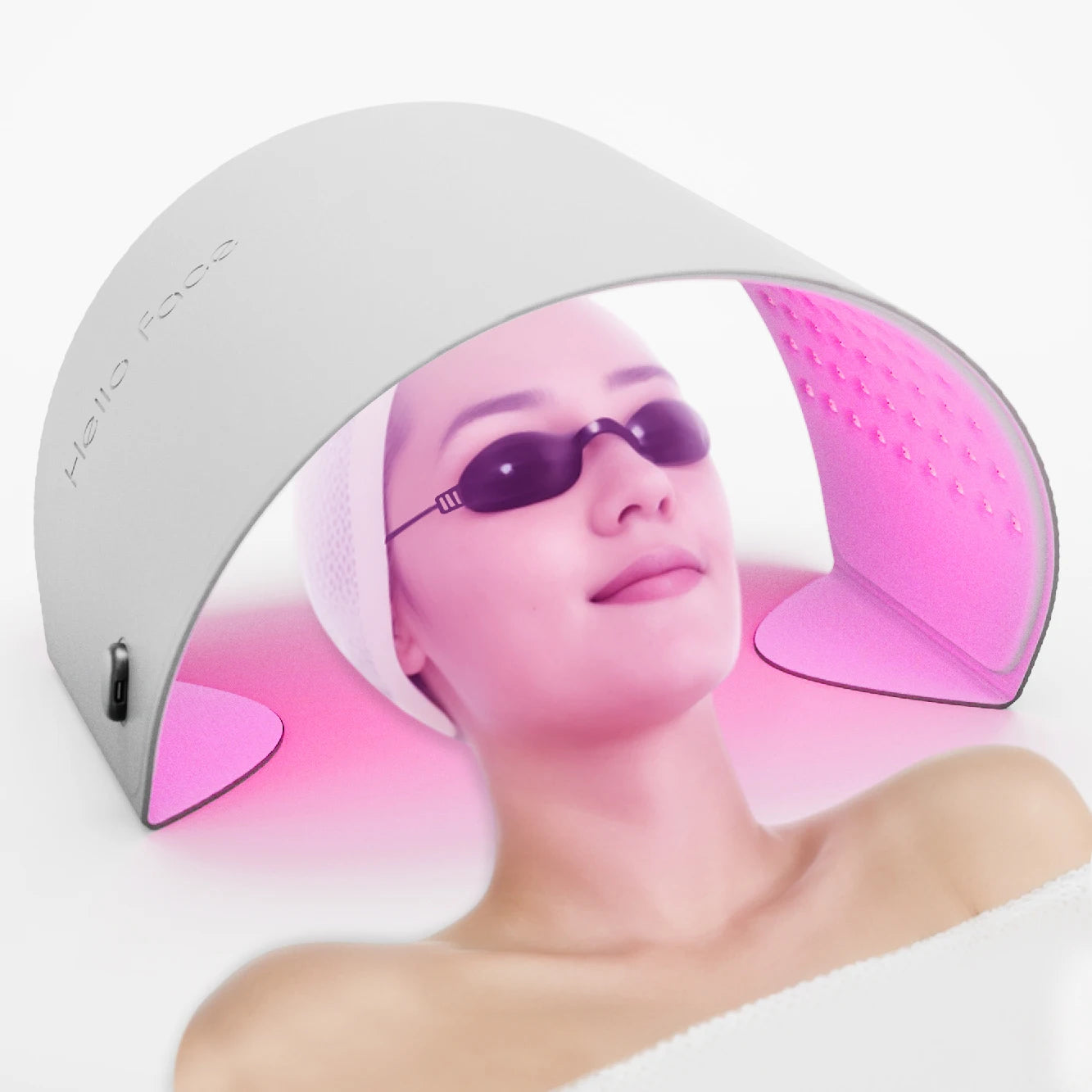 A woman wearing goggles lies under an arched, white Hello Face LED therapy device emitting a vibrant pink light.