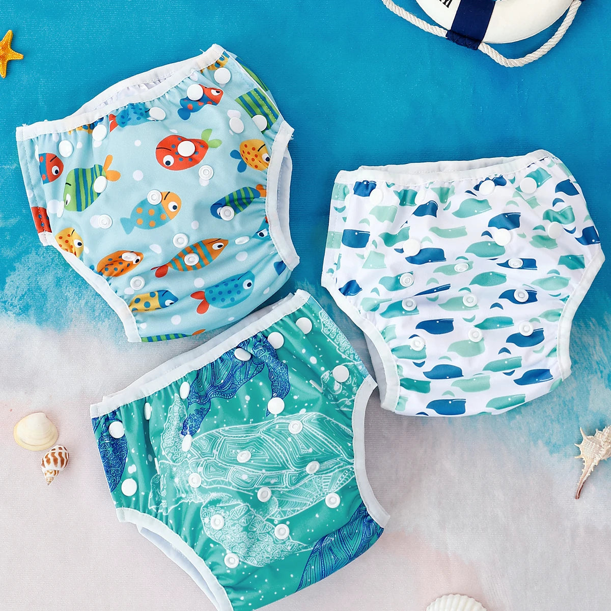 Three adjustable baby swim diapers featuring fish, whale, and sea turtle patterns on a blue beach-themed background.