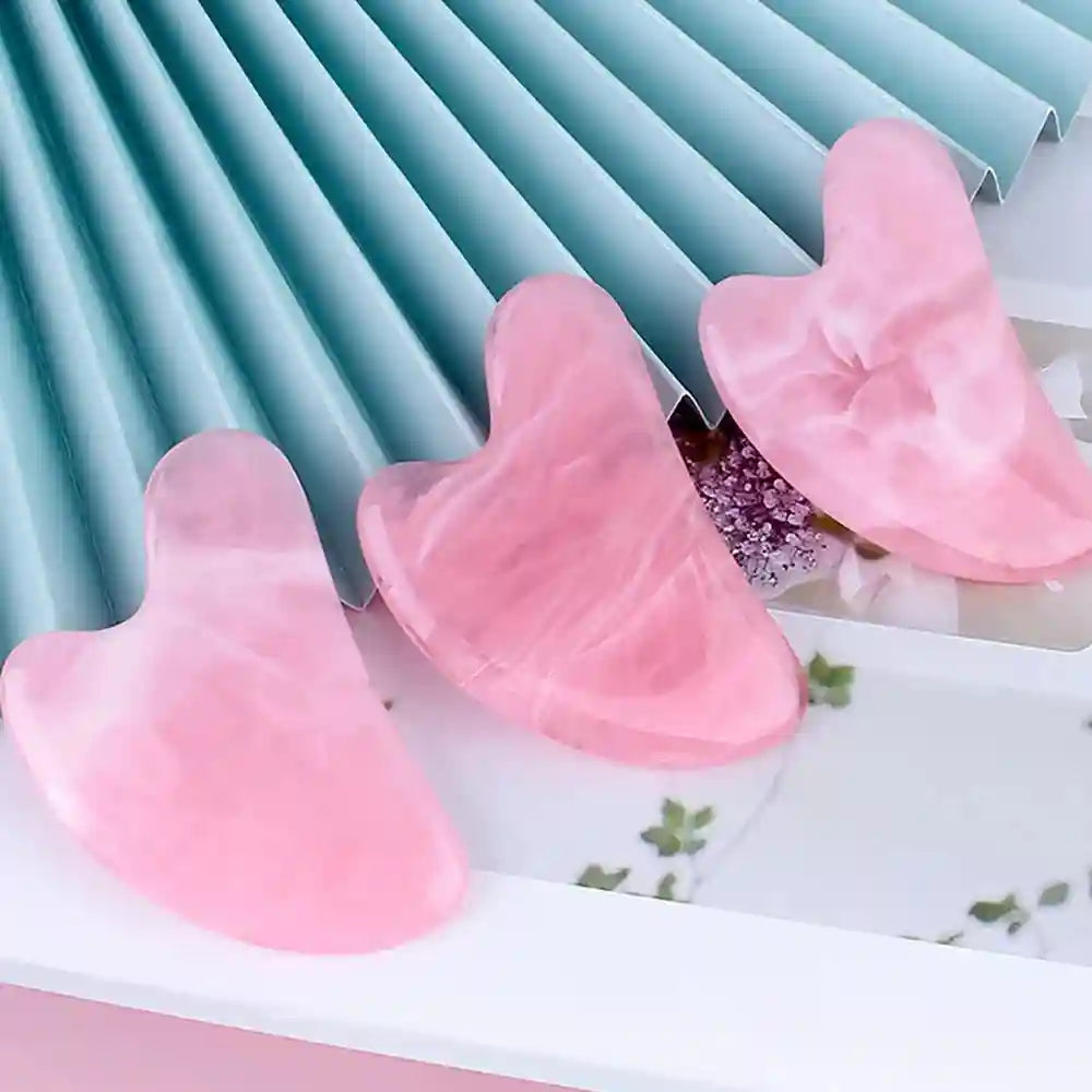 Three heart-shaped, pink marbled resin gua sha massage tools displayed against a blue pleated background.