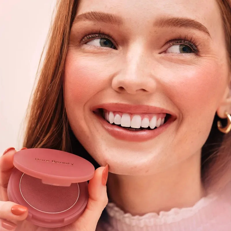 Woman holding a Rare Beauty powder blush in a soft pink compact with a smooth, matte finish and natural-looking pigment.