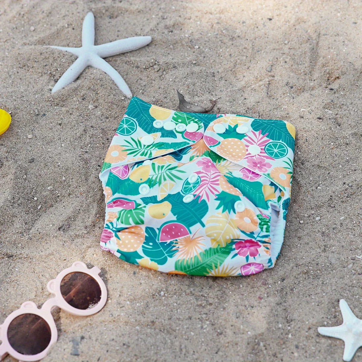 Reusable cloth baby diaper with colorful tropical fruit and leaf print on a sandy beach background.