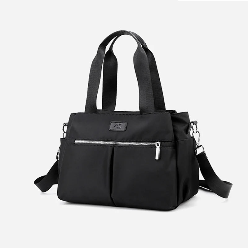 Minimalist black canvas satchel handbag with top handles, silver front zipper, and adjustable crossbody shoulder strap.