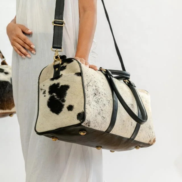 Unisex cow hair-on-hide weekender duffel bag with black leather trim, shoulder strap, and brass hardware trunk style.