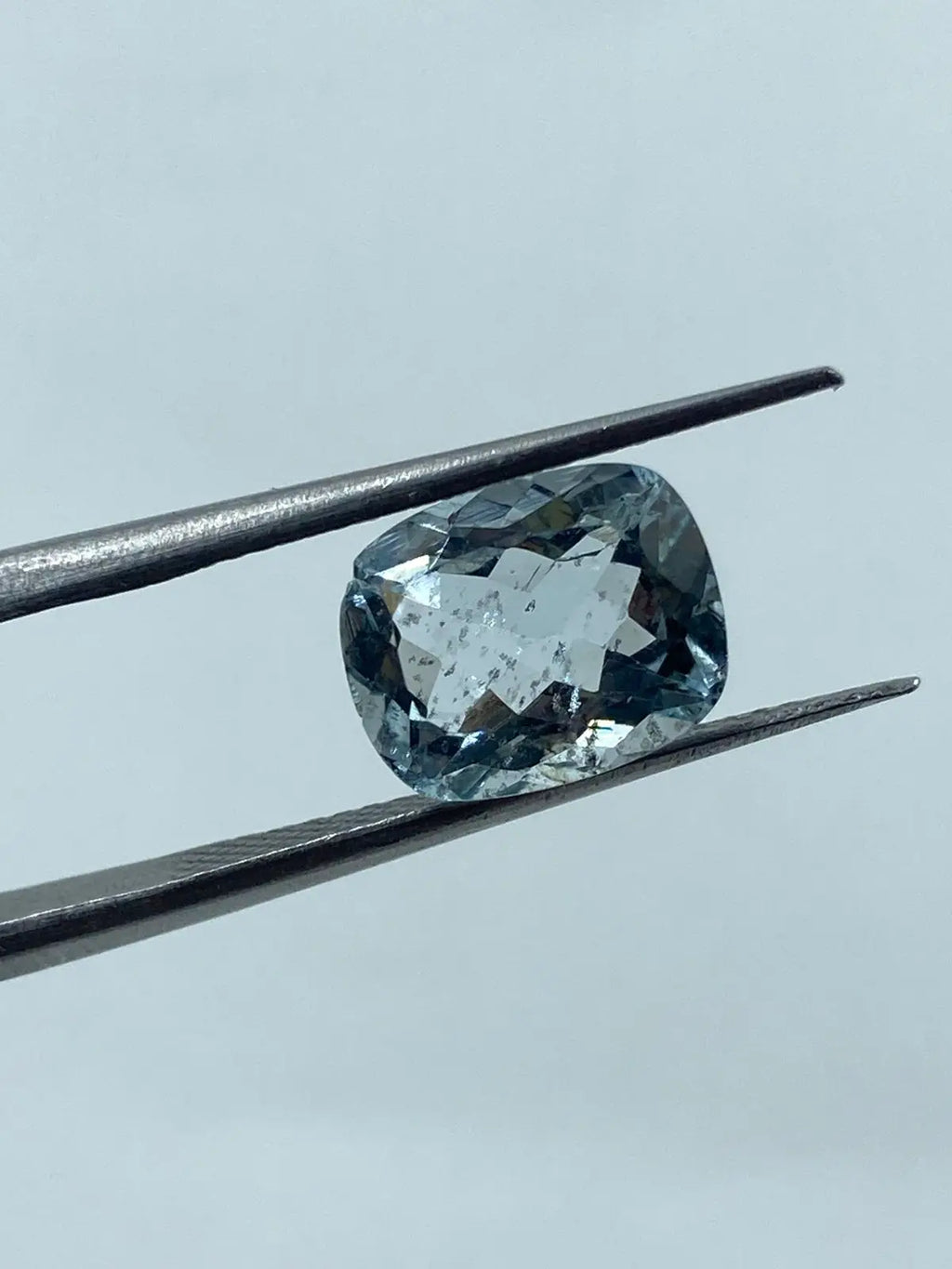 Sky-blue cushion-cut gemstone between metal tweezers, faceted