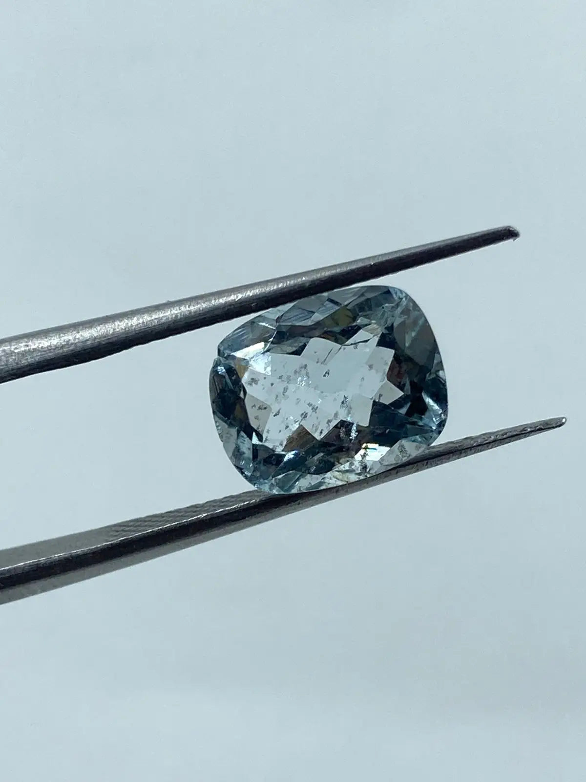 Sky-blue cushion-cut gemstone between metal tweezers, faceted