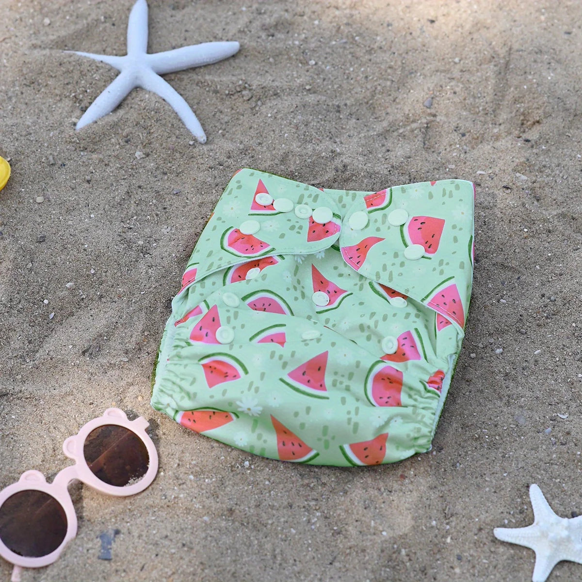 Watermelon print reusable baby cloth diaper with adjustable snaps on beach sand background.
