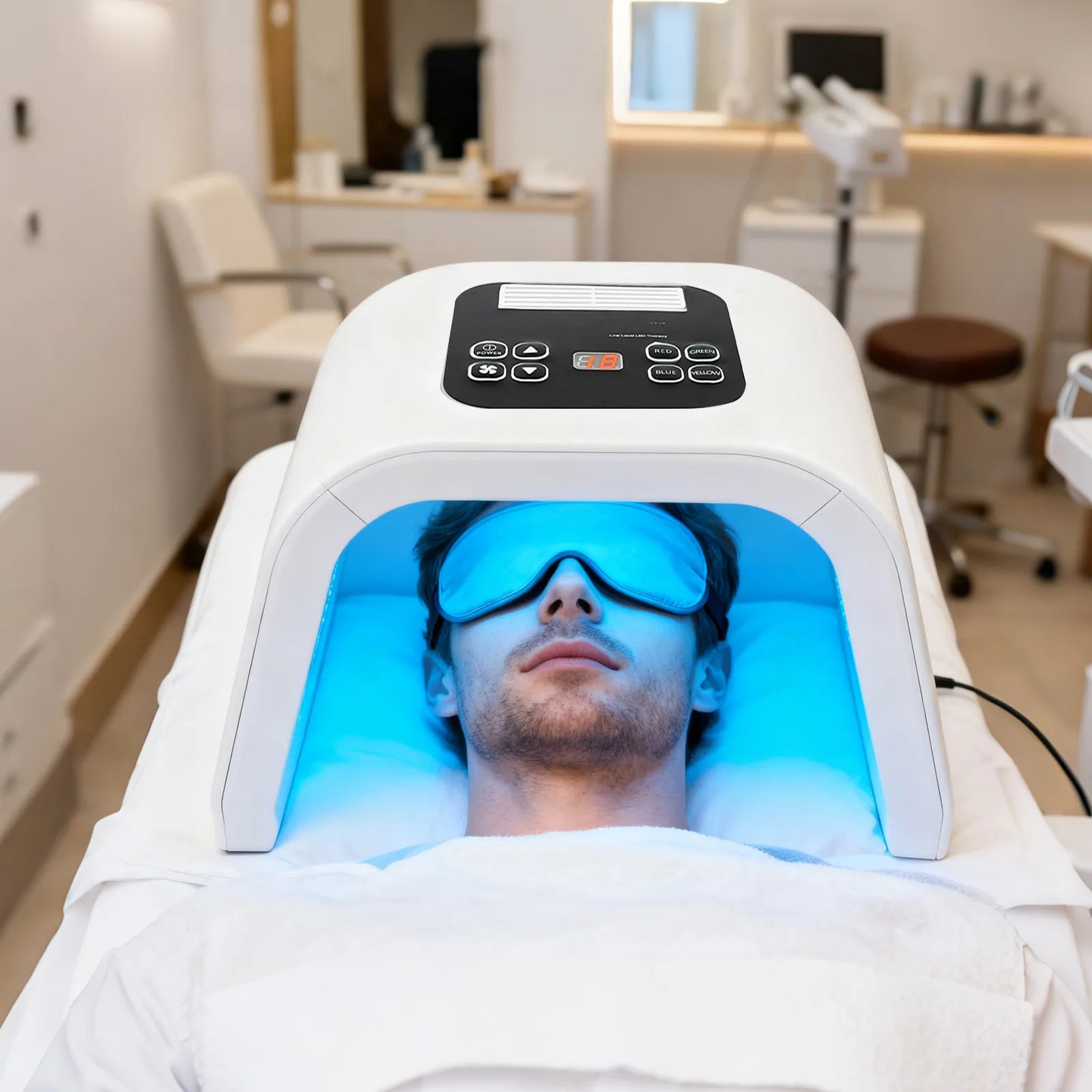 A man lying under a white, arched LED light therapy machine, wearing blue eye protection in a spa setting.
