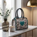Genuine leather mini trunk bag with teal heart, floral pattern, and black handles. Luxury weekender duffle style.