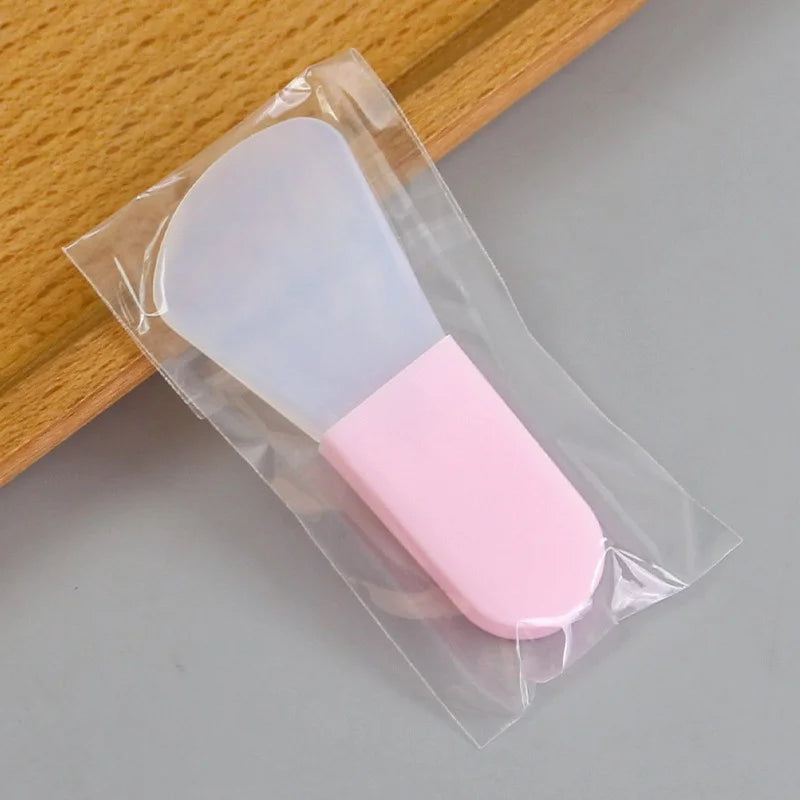 A silicone facial mask applicator with a translucent white flexible head and a solid pastel pink handle in plastic wrap.