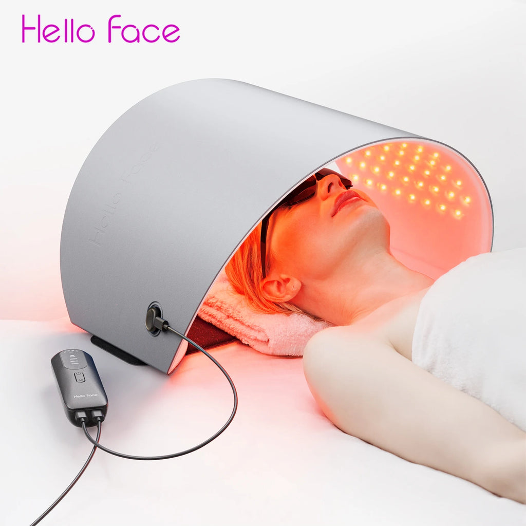 A person lying under a silver, arched Hello Face LED therapy panel emitting red light for skin rejuvenation.