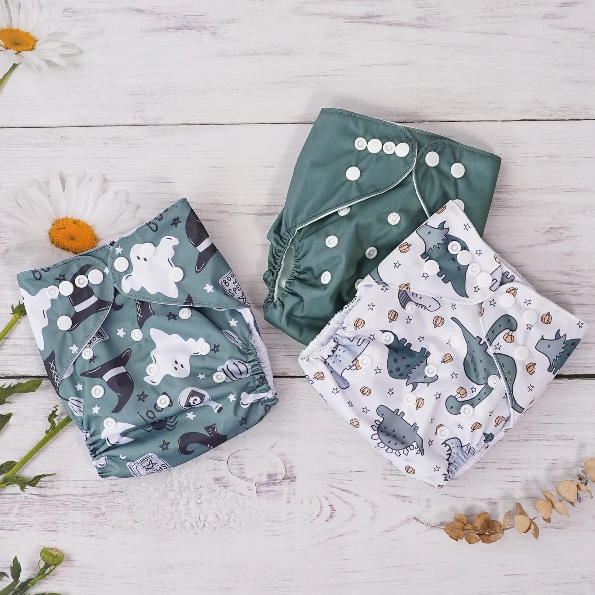 Three cloth diapers with snap closures, featuring green solid, ghost/witch, and dinosaur baby care prints.