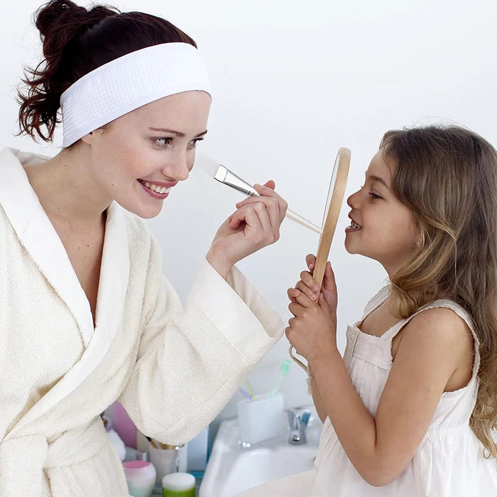 A woman in a robe uses a clear-handled facial mask brush to playfully apply product to a young girl holding a mirror.