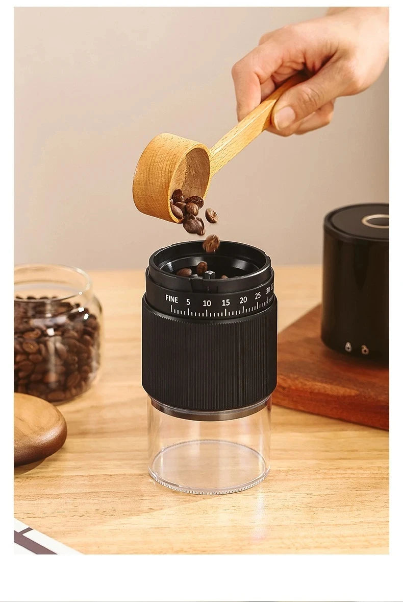 Hand pouring coffee beans from a wooden scoop into a black, compact manual coffee grinder with clear base.