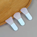 Three silicone face mask applicators with opaque white fan-shaped heads and light blue handles on a wooden tray.
