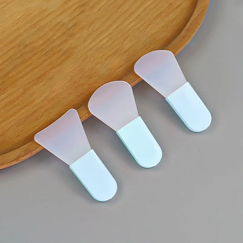 Three silicone face mask applicators with opaque white fan-shaped heads and light blue handles on a wooden tray.