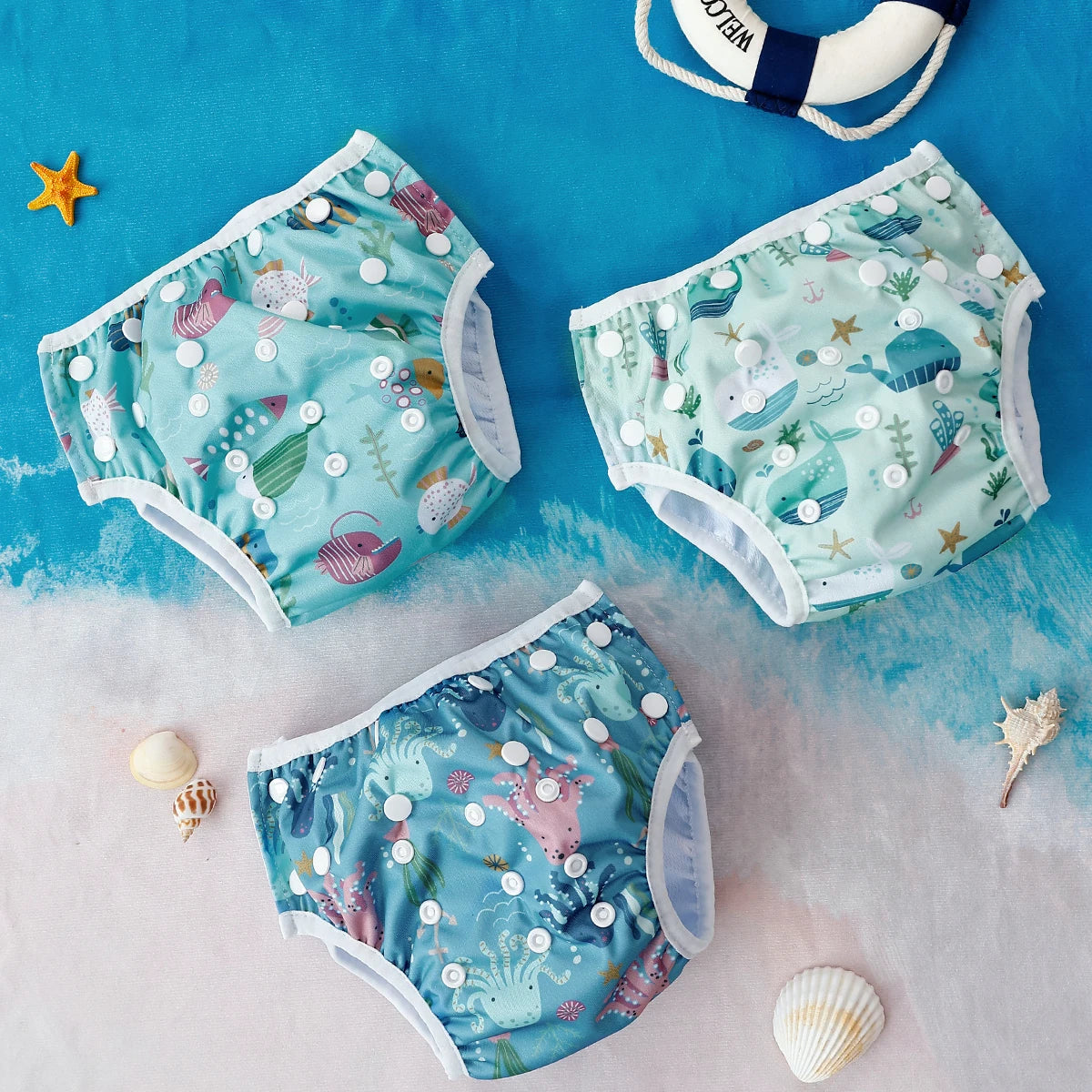 Three reusable baby swim diapers with ocean-themed whale, fish, and octopus prints on a beach background.