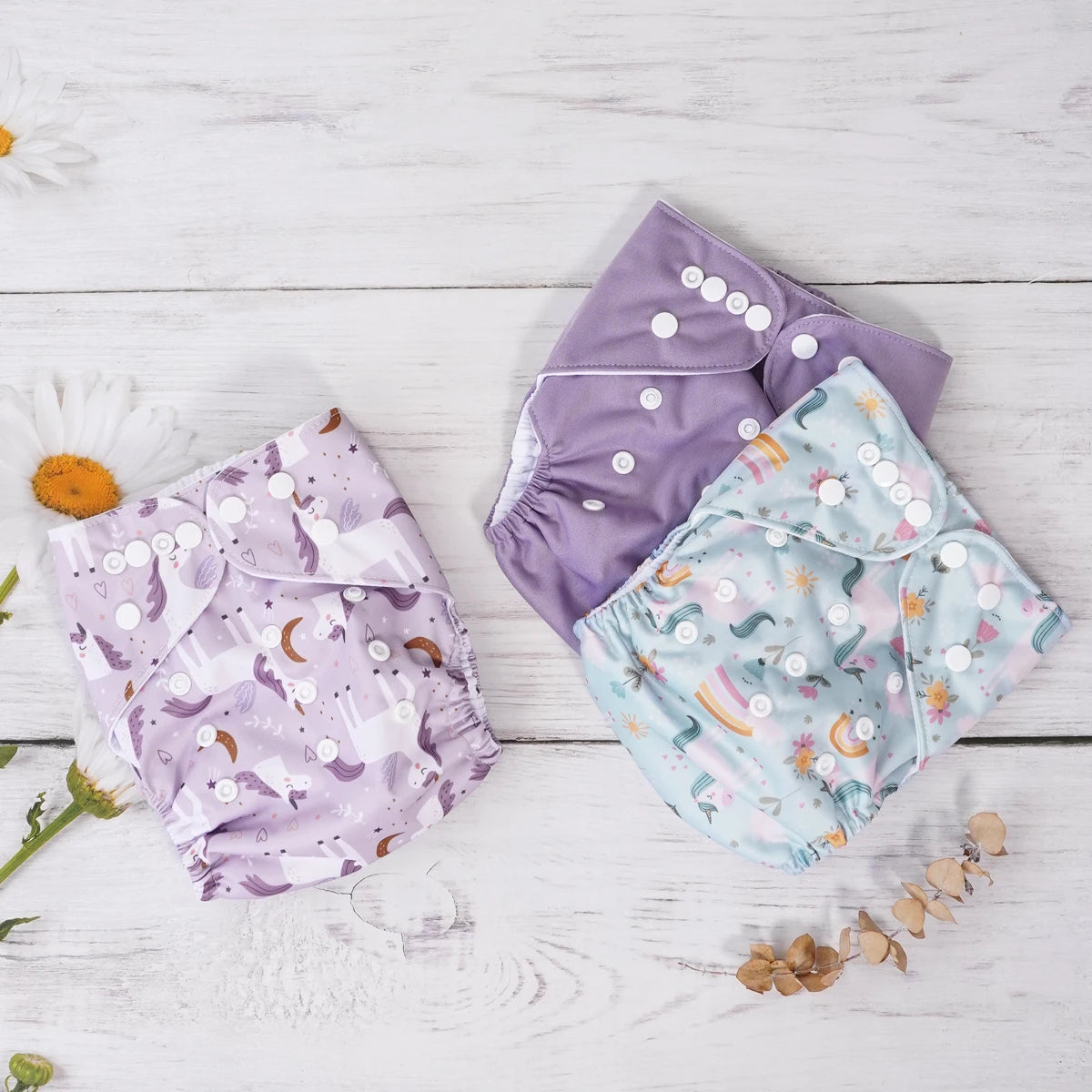 Three adjustable cloth baby diapers featuring unicorn, floral, and solid purple designs on a white wooden background.
