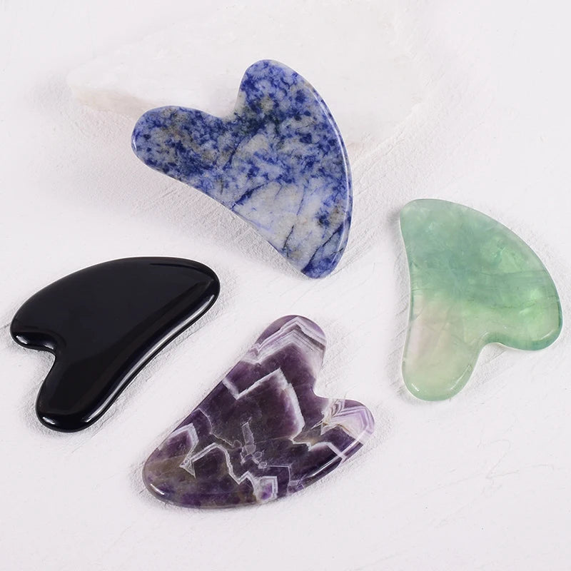 Four heart-shaped stone Gua Sha facial massagers in blue, black, purple, and green on a white background.