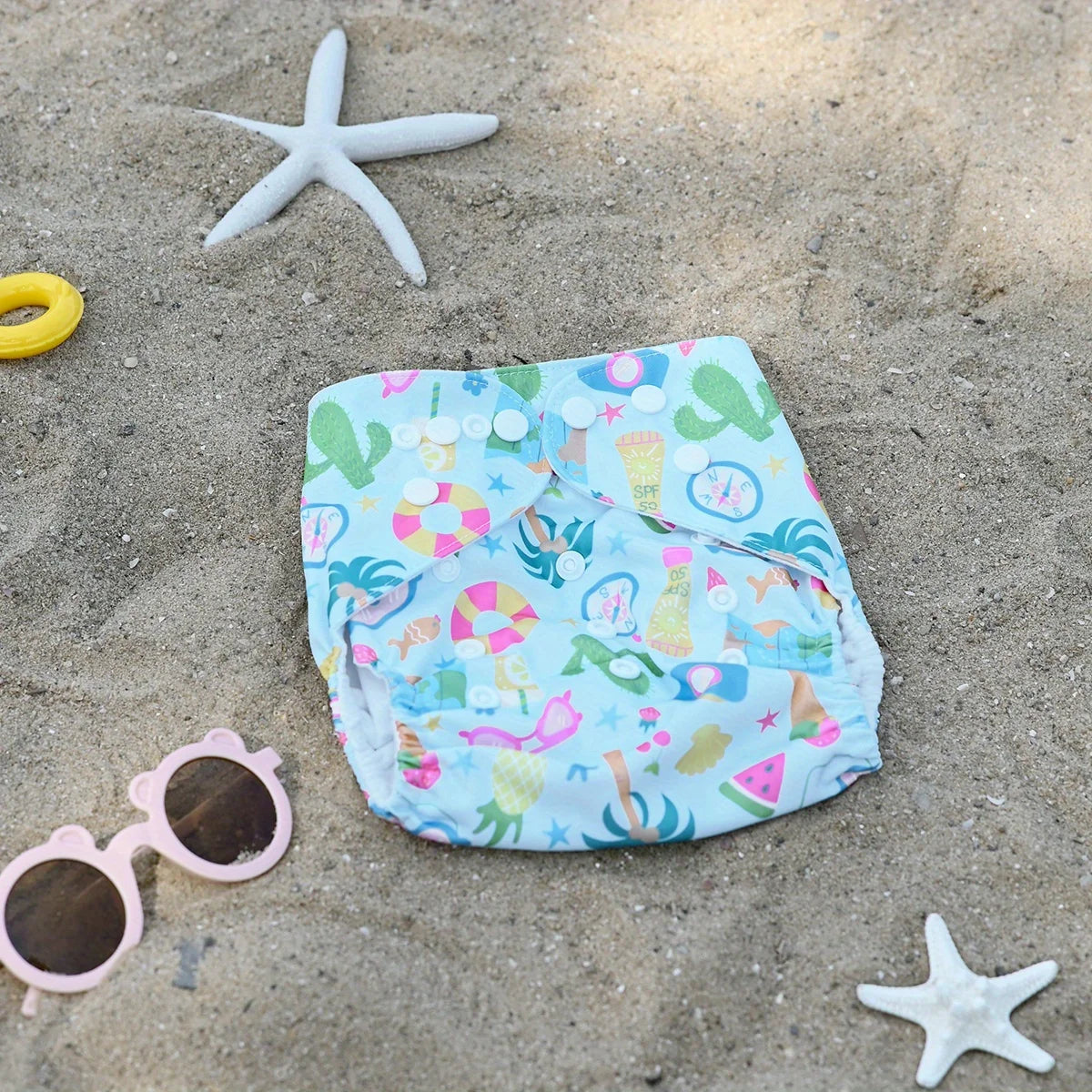 Reusable cloth swim diaper with summer beach print, featuring cacti, tropical fruits, and SPF sunscreen icons.