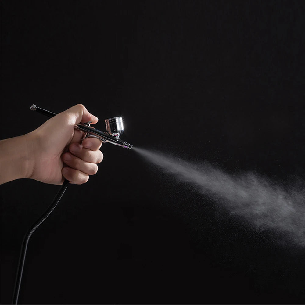 A hand holding a metallic silver airbrush gun, spraying a fine mist against a plain black background.