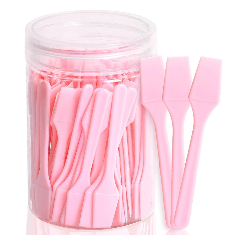 A set of 100 small, pink plastic cosmetic spatulas for makeup or skincare, stored in a clear plastic cylindrical jar.