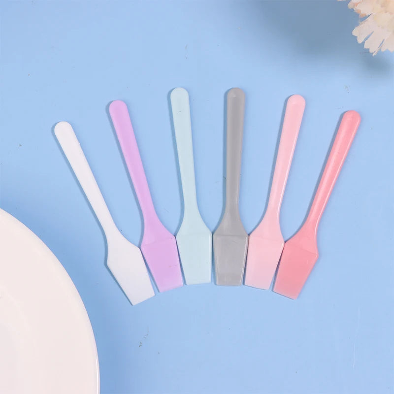 Six small, multi-colored plastic cosmetic spatulas fanned out on a light blue background.