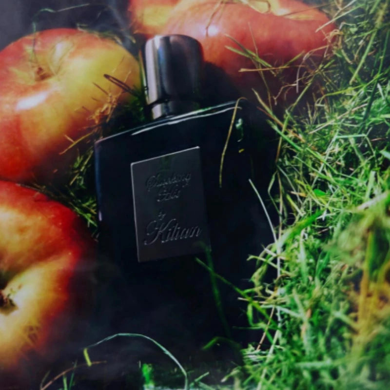 Black rectangular glass perfume bottle with a silver label, nestled in grass among fresh red apples.