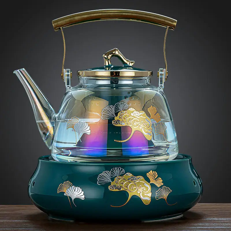 Elegant glass tea kettle with gold trim, ginkgo leaf pattern, and teal ceramic base on a wooden table.