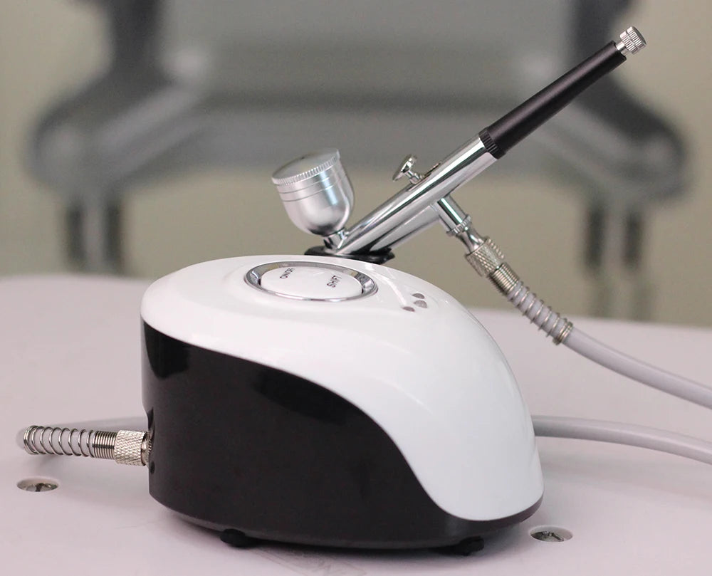 A black and white portable airbrush machine with a silver metallic spray gun attached by a hose.