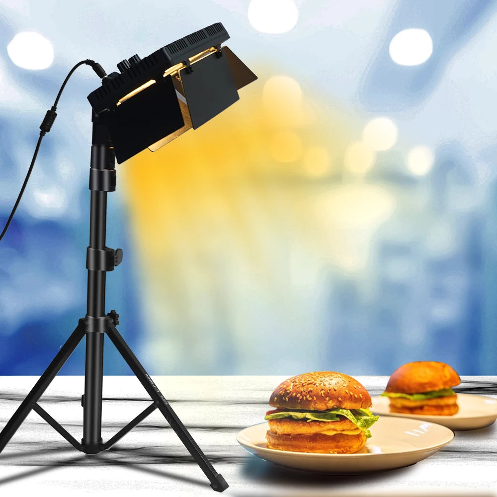 Black rectangular LED video light with adjustable metal barn doors on a tripod stand, illuminating plates of burgers.