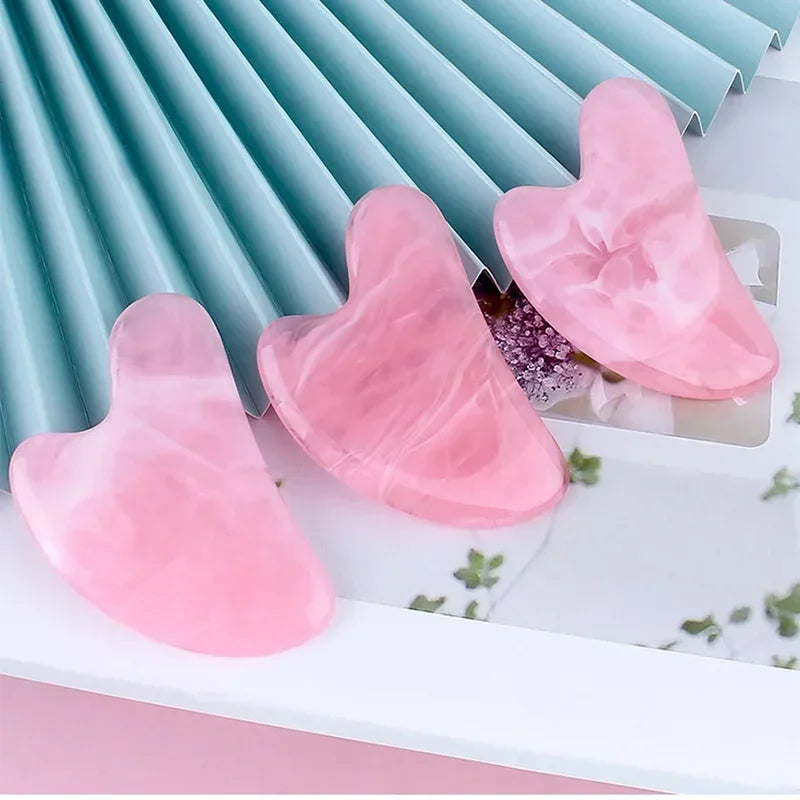 Three heart-shaped, pink marbled resin gua sha facial massage tools resting on a light blue pleated background.
