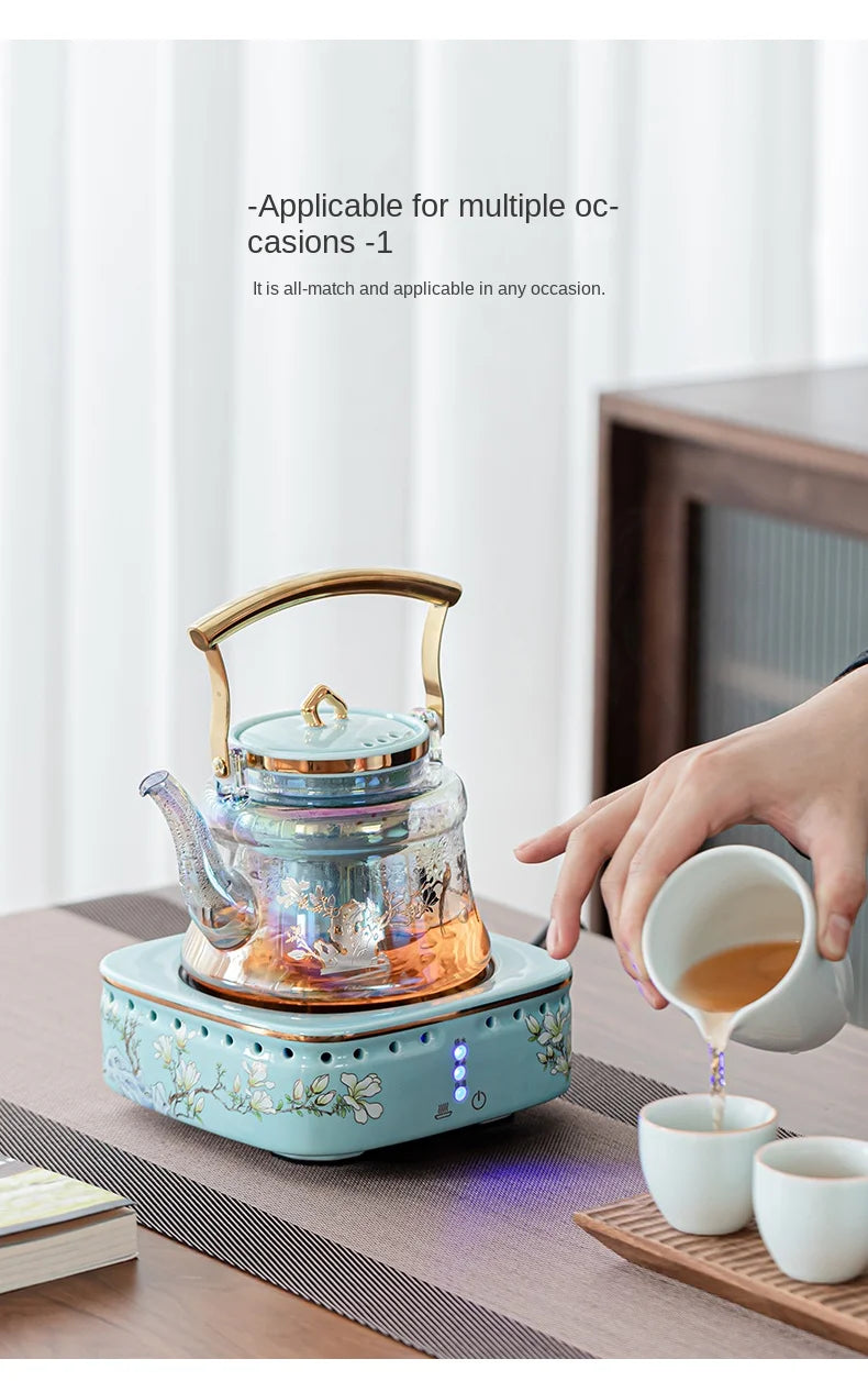 Iridescent glass tea kettle with gold handle on a floral ceramic electric heating base for brewing tea.