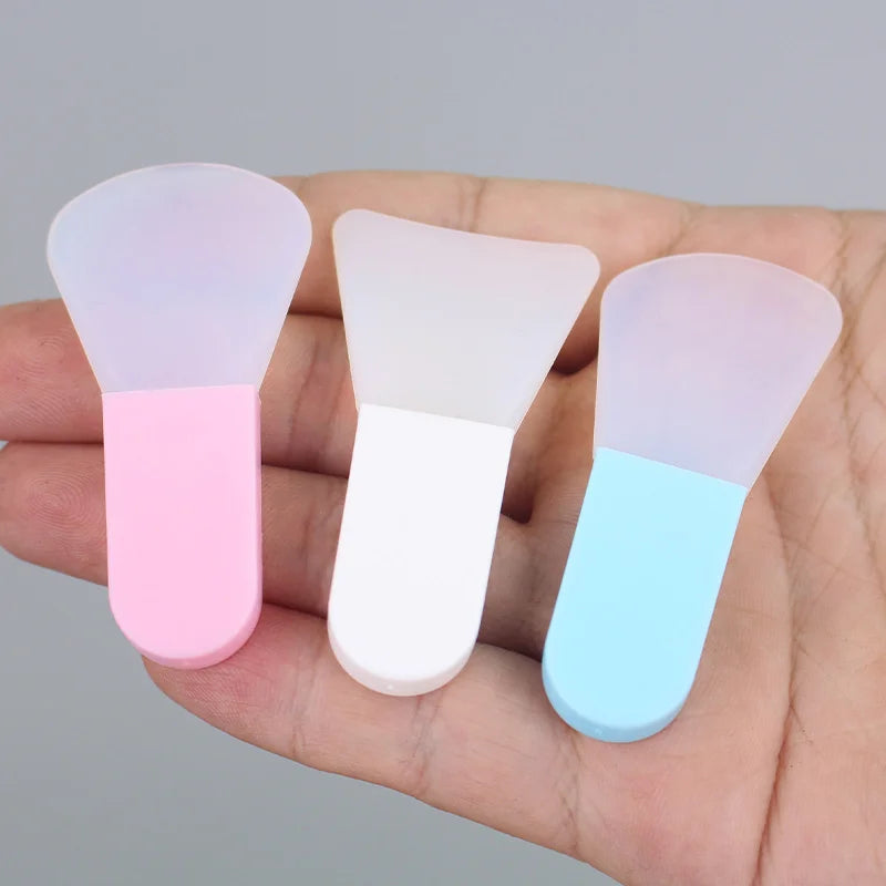 Three small, portable silicone face mask applicators with pink, white, and blue handles held in a palm.
