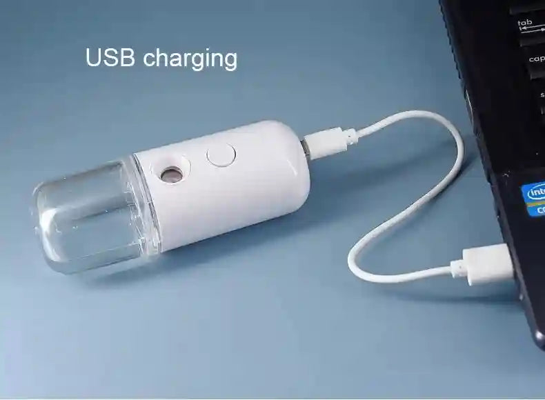 Portable white nano facial mister sprayer for hydrating skincare and eyelashes charging via USB with a laptop.