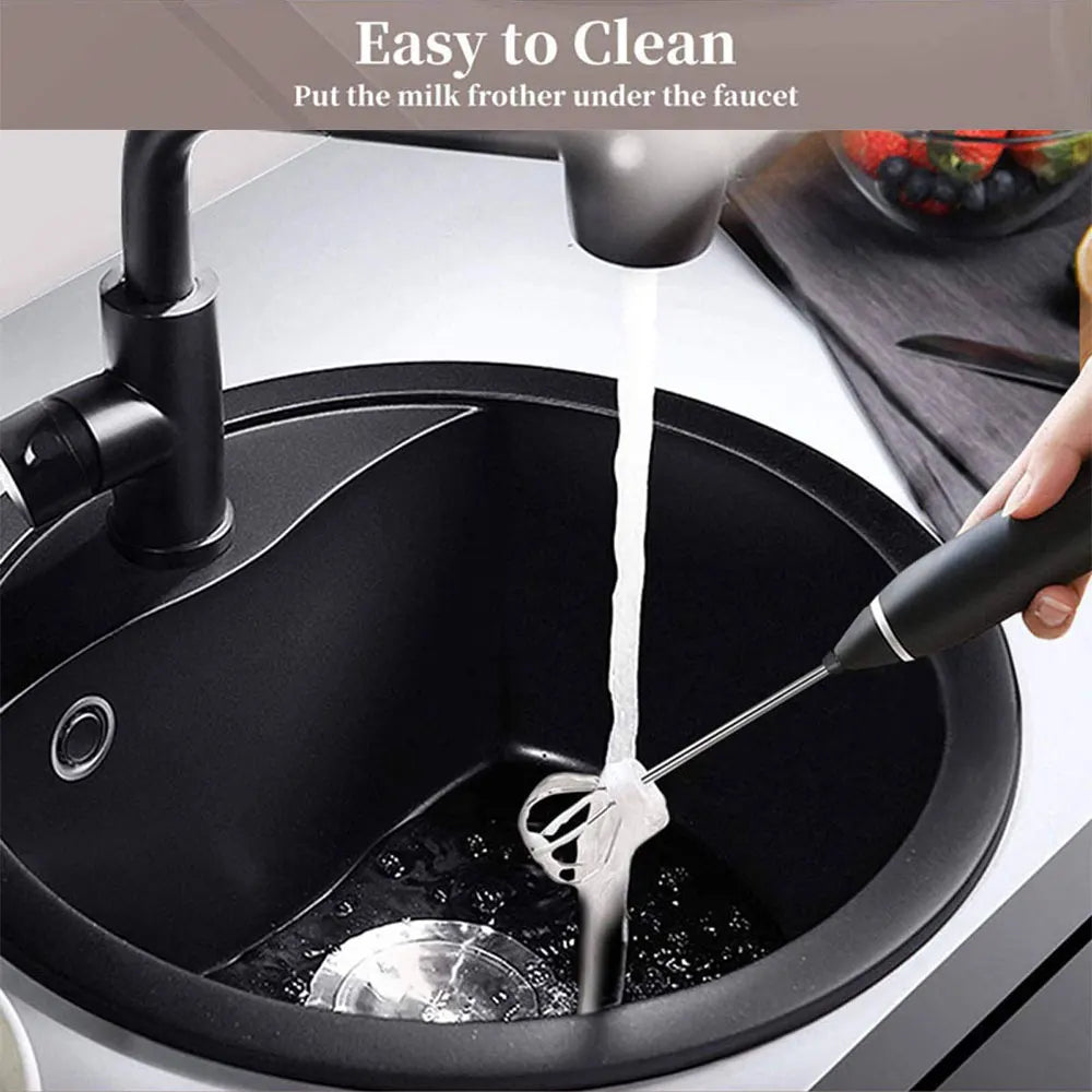 Handheld electric milk frother with black handle and stainless steel whisk being rinsed under a kitchen faucet.