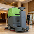 Commercial floor scrubber machine with green tank and steering wheel for large indoor lobby and commercial floor cleaning.