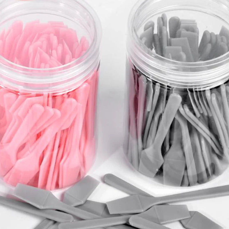 Two clear jars filled with small, pink and grey plastic cosmetic spatulas for applying creams and masks.