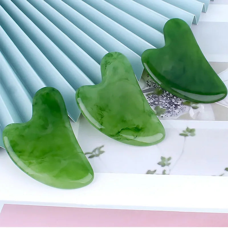 Three smooth, heart-shaped green resin Gua Sha facial massage tools on a light surface with a blue pleated background.