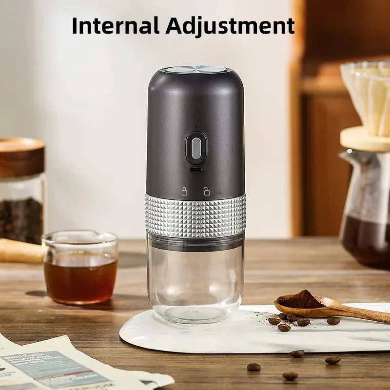Portable electric coffee grinder in matte black and silver with a clear plastic grounds container on a wooden table.