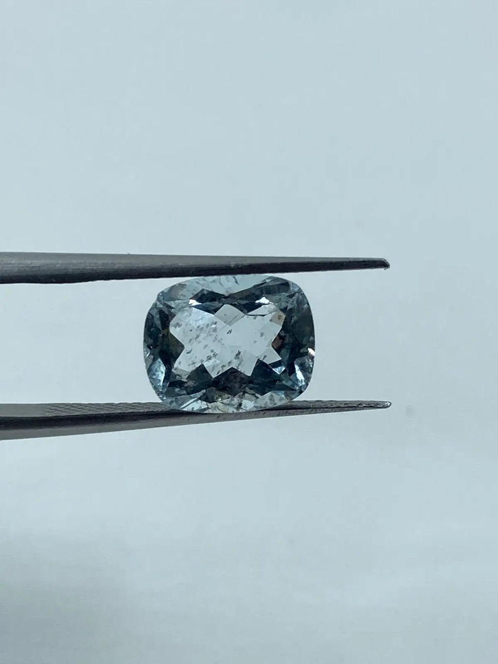 Loose cushion-cut aquamarine gemstone, sky blue, held between tweezers, frontal view