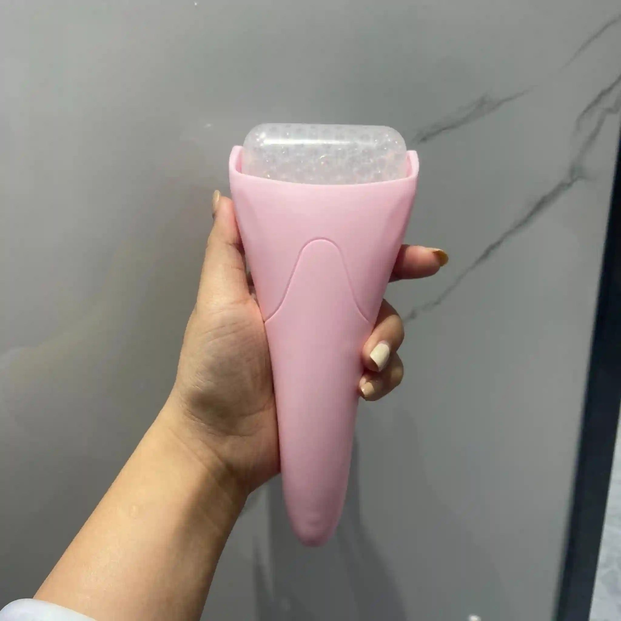 A hand holds a pink, tapered facial ice roller with a translucent, bubbly textured cooling head.