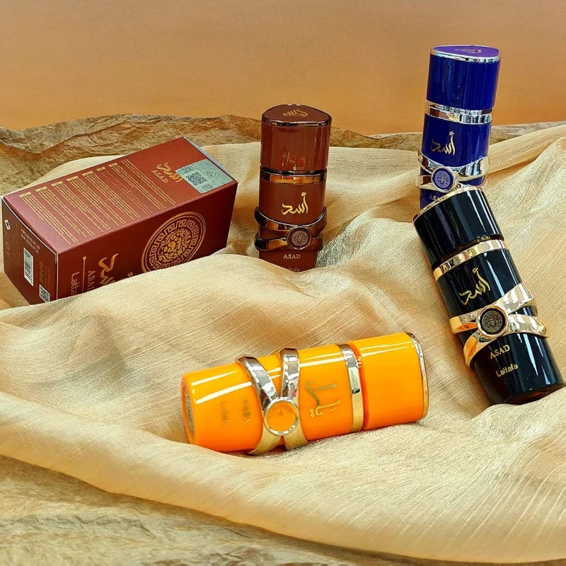 Asad perfume collection: four cylindrical bottles in orange, brown, black, and blue with gold accents on a gold fabric.