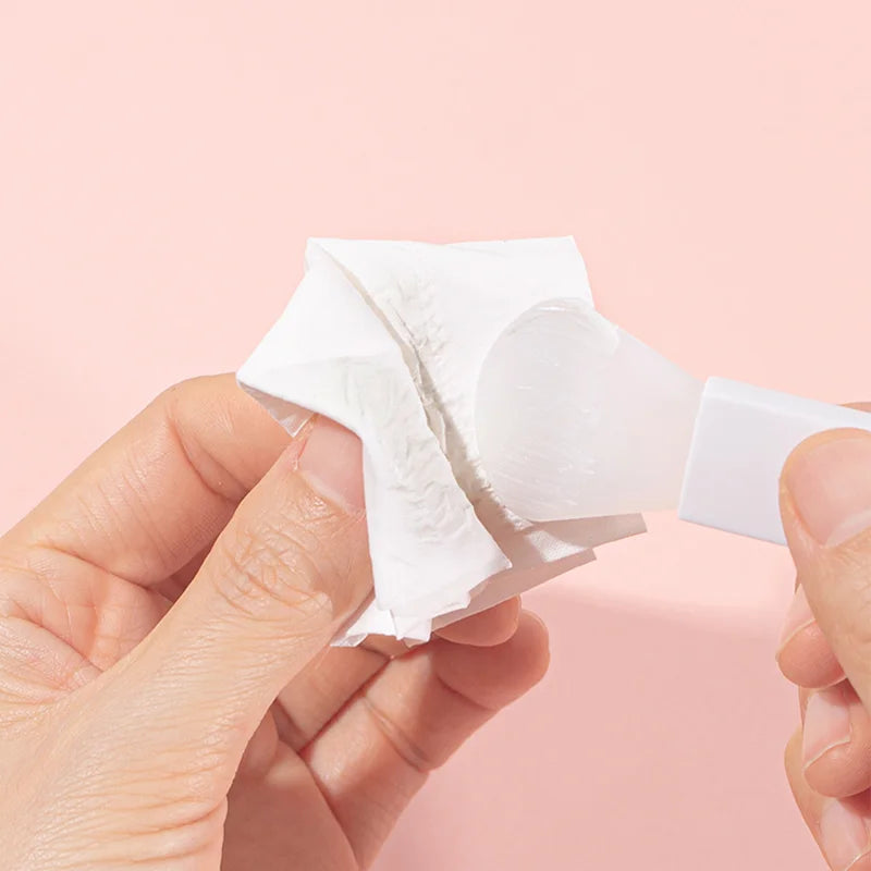 Hands holding a white silicone facial mask applicator brush, cleaning it with a white tissue on a pale pink background.