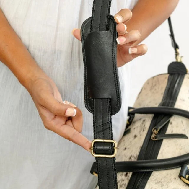 Black leather shoulder strap with padded cushion on a cow hair-on-hide weekender travel duffel bag.