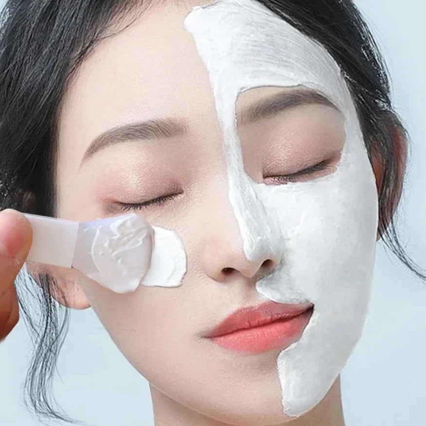 A person applying a white mud face mask with a flexible, flat-tipped white silicone applicator tool.