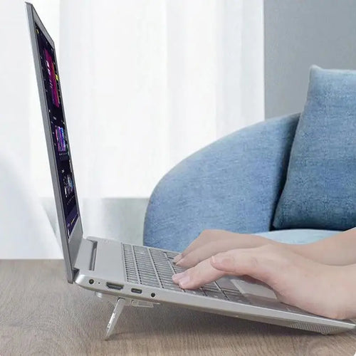 Silver laptop on a compact, angled foldable metal stand, being used on a wooden desk with a sofa in the background.