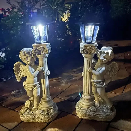 Two off-white resin cherub angel statues holding lit solar lantern pillars for outdoor garden lighting.